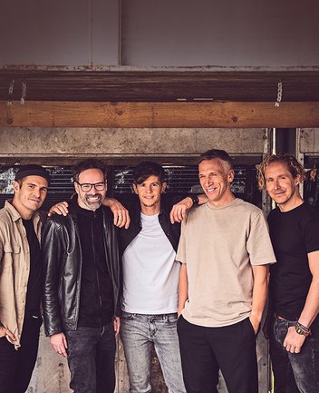 Die Band Kasalla ist als Gruppe von fünf Männern zu sehen, die eng nebeneinander stehen und sich teilweise die Arme um die Schultern legen. Sie tragen lässige Kleidung wie T-Shirts, Hemden und Lederjacke und blicken entspannt in die Kamera. Im Hintergrund ist eine rustikale Wand mit Holz- und Metallelementen zu erkennen, die eine warme, ungezwungene Atmosphäre vermittelt.
