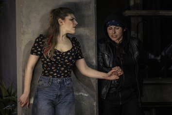 Amenaide und Tancredi stehen in einer angespannten Szene nebeneinander. Amenaide lehnt an einer Wand und blickt besorgt zur Seite, während Tancredi, dunkel gekleidet mit Lederjacke und Kopftuch, aus einem Schattenbereich hervortritt. Die beiden halten sich an den Händen, was ihre enge Verbindung unterstreicht. Die reduzierte, moderne Bühnenausstattung und das gedämpfte Licht verleihen der Szene aus Rossinis Oper Tancredi an der Oper Köln eine intime und dramatische Atmosphäre.