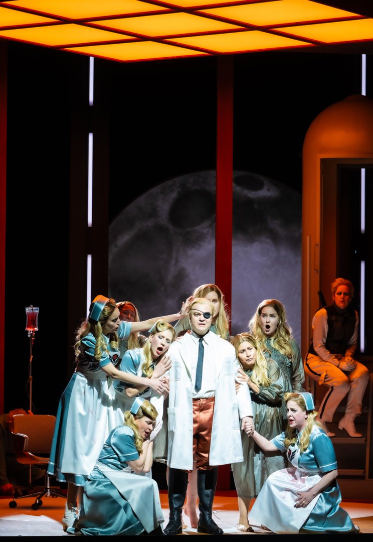 Eine stilisierte Opernszene aus Wagners Die Walküre zeigt Wotan im Zentrum, umgeben von den Walküren, während Brünnhilde im Hintergrund in einer raketenartigen Kapsel sitzt.
Wotan steht frontal im Mittelpunkt der Bühne, selbstbewusst und nahezu regungslos. Er trägt ein modernes Kostüm: ein weißer Mantel über Hemd und Krawatte, dazu glänzende, rostfarbene Shorts und schwarze Stiefel. Eine Augenklappe verleiht ihm ein markantes, autoritäres Erscheinungsbild.
Um ihn herum gruppieren sich die Walküren in hellblauen und weißen, retro anmutenden Uniformen, die an Krankenschwestern erinnern. Einige knien oder beugen sich zu ihm, berühren seine Arme und Schultern oder schauen ihn mit intensiven, emotional aufgeladenen Gesichtsausdrücken an – zwischen Verehrung, Sorge und dramatischer Erregung.
Im rechten Hintergrund sitzt Brünnhilde in einer kapselartigen, raketenähnlichen Struktur mit Tür und kleiner Treppe. Ihre Körperhaltung wirkt angespannt und isoliert, als sei sie von der Gruppe getrennt oder in einer eigenen Situation gefangen.
Die Bühne ist stark stilisiert: Über den Figuren spannt sich eine leuchtend orangefarbene Decke aus geometrischen Lichtflächen, die den Raum in warmes, künstliches Licht taucht. Dahinter ist ein großer, diffuser Mond sichtbar, der einen Kontrast zwischen futuristischer Innenarchitektur und mythischer Bildwelt schafft. Insgesamt entsteht eine Mischung aus Science-Fiction-Ästhetik, Ritual und emotional aufgeladener Gruppenszene.