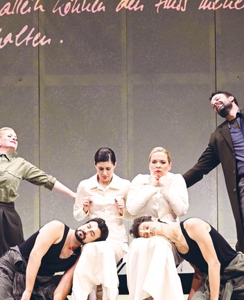 Six performers from Così fan tutte on stage: In the center sit two women (Fiordiligi and Dorabella) in elegant, light, cream-colored, high-necked dresses. At their feet lie two men (Ferrando and Guglielmo) in dark tank tops and casual trousers, with a slightly disheveled, modern appearance. On the left stands a woman (Despina) wearing an olive-green shirt and a dark skirt; on the right, a man (Don Alfonso) in a dark coat over a shirt and trousers. The costumes combine classical and modern elements, creating a stylized, timeless impression.