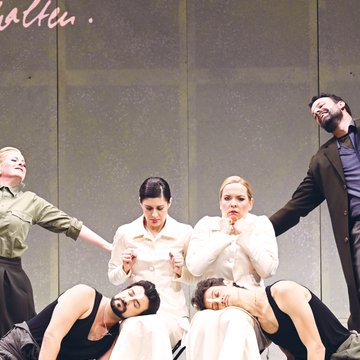 Six performers from Così fan tutte on stage: In the center sit two women (Fiordiligi and Dorabella) in elegant, light, cream-colored, high-necked dresses. At their feet lie two men (Ferrando and Guglielmo) in dark tank tops and casual trousers, with a slightly disheveled, modern appearance. On the left stands a woman (Despina) wearing an olive-green shirt and a dark skirt; on the right, a man (Don Alfonso) in a dark coat over a shirt and trousers. The costumes combine classical and modern elements, creating a stylized, timeless impression.