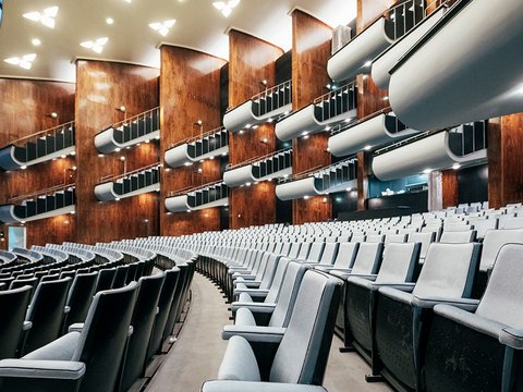 Weitwinkelblick in den leeren Zuschauerraum der Oper Köln am Offenbachplatz: In sanft geschwungenen Reihen erstrecken sich zahlreiche hellblaue, gepolsterte Sitze nach vorne zur Bühne hin. Entlang der Seiten und Rückwand befinden sich mehrere übereinanderliegende Ränge mit Balkonen, deren Brüstungen in hellen, geschwungenen Formen gestaltet sind. Die Wände sind mit warmen, rötlich-braunen Holzpaneelen verkleidet, die dem Raum eine elegante, moderne Atmosphäre verleihen. Kleine, punktuelle Leuchten setzen Akzente entlang der Ränge. Die hohe Decke ist hell und mit gleichmäßig verteilten, geometrisch angeordneten Leuchtelementen versehen, die den Raum weich ausleuchten. Insgesamt vermittelt der Saal einen großzügigen, modernen und architektonisch markanten Eindruck.