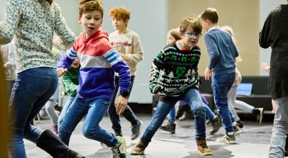 Eine Schulklasse bewegt sich aktiv durch einen großen Innenraum. Mehrere Kinder laufen, springen und reagieren aufeinander in einer spielerischen Gruppenübung. Im Vordergrund sind zwei Kinder in dynamischer Bewegung zu sehen, während weitere Schüler*innen im Hintergrund teilnehmen. Die Szene vermittelt Energie, Teamarbeit und gemeinsames Lernen durch Bewegung.