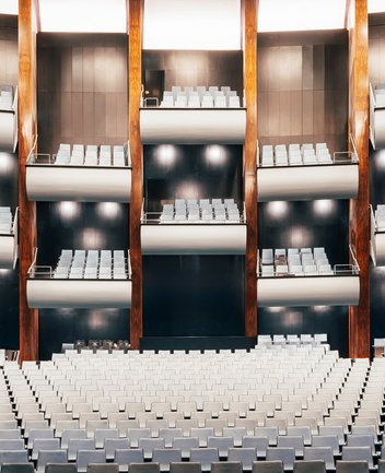 Weitwinkelansicht des großen Zuschauerraums der Oper Köln am Offenbachplatz mit mehreren übereinanderliegenden Rängen. Die hellen Sitzreihen füllen den Raum gleichmäßig, während vertikale Holzpaneele und dunkle Wandflächen die Architektur strukturieren.