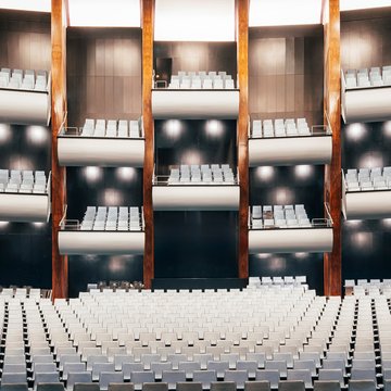 Weitwinkelansicht des großen Zuschauerraums der Oper Köln am Offenbachplatz mit mehreren übereinanderliegenden Rängen. Die hellen Sitzreihen füllen den Raum gleichmäßig, während vertikale Holzpaneele und dunkle Wandflächen die Architektur strukturieren.