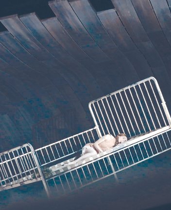 Eine Darstellerin als Nixe aus Rusalka von Antonín Dvořák liegt seitlich eingerollt auf einem schräg stehenden, weißen Metallbettgestell. Sie trägt ein helles, ärmelloses Kostüm in zarten Farbtönen. Die Bühne ist in kühlen, bläulichen Lichtstimmungen gehalten, im Hintergrund erheben sich geschwungene, wellenartige Strukturen, die eine Unterwasserwelt andeuten. Die Szene wirkt ruhig, traumartig und leicht surreal.