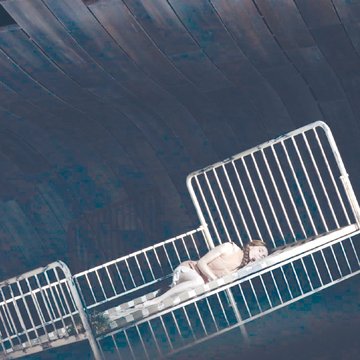 A female performer as the Water Nymph from Rusalka by Antonín Dvořák lies curled on her side on a tilted, white metal bed frame. She wears a light, sleeveless costume in soft tones. The stage is dimly lit with cool, bluish hues, and curved, wave-like structures rise in the background, evoking an underwater atmosphere. The scene appears quiet, dreamlike, and slightly surreal.