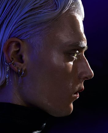 Nahaufnahme eines jungen Mannes mit cineastischer Bildwirkung und warmer Lichtstimmung: Der junge Mann ist im Profil nach rechts gewandt und blickt konzentriert in die Ferne, der Ausdruck wirkt wachsam und entschlossen. Das kurze, silbrig-blonde Haar ist nach hinten gekämmt. Mehrere Ohrringe und Ketten im Ohr verleihen der Erscheinung eine moderne, leicht rebellische, cyberpunkartige Note. Die Haut glänzt im Bühnenlicht, wodurch das Gesicht transparent wirkt und die Konturen des Gesichts betont werden. Die Beleuchtung ist kontrastreich und in kühlen Blau- und Violetttönen gehalten, was eine intensive, dramatische Atmosphäre erzeugt. Der dunkle Hintergrund verstärkt die Fokussierung auf das Gesicht und unterstreicht die kraftvolle, zeitgenössische Interpretation der Figur des Siegfried aus Richard Wagners Oper Siegfried.