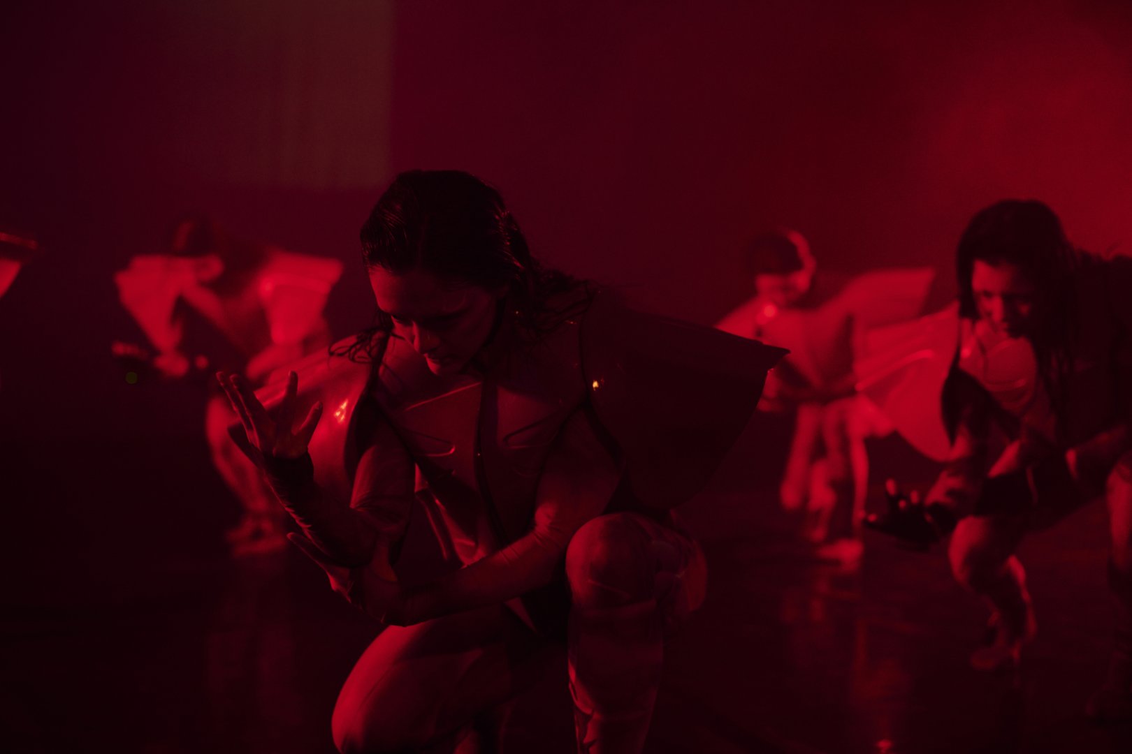 Mehrere Tänzerinnen in futuristisch anmutenden Kostümen bewegen sich in einer intensiv rot beleuchteten Bühne. Im Vordergrund kniet eine Person, den Oberkörper leicht nach vorne geneigt, und betrachtet konzentriert ihre erhobene Hand. Weitere Performerinnen sind im Hintergrund in ähnlichen, fließenden Bewegungen zu sehen. Die Szene wirkt konzentriert, technisch und spannungsgeladen.
