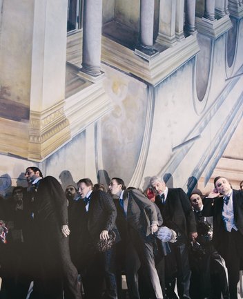 Szene aus Rigoletto von Giuseppe Verdi: Eine Gruppe elegant gekleideter Männer in dunklen Anzügen und Fräcken steht dicht gedrängt vor einer klassizistischen Architekturkulisse mit Säulen und Balustraden; ihre Körperhaltung wirkt geneigt und gespannt, als würden sie gemeinsam auf etwas reagieren.