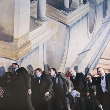 Scene from Rigoletto by Giuseppe Verdi: A group of elegantly dressed men in dark suits and tailcoats stand closely packed in front of a neoclassical architectural backdrop with columns and balustrades; their bodies lean forward, tense, as if reacting to something together.