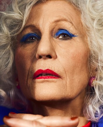 Nahaufnahme einer alten Dame mit cineastischer Bildwirkung und warmer Lichtstimmung: Die ältere Frau blickt frontal in die Kamera, ihr Ausdruck wirkt entschlossen und zugleich herausfordernd. Ihr silbergraues, lockiges Haar umrahmt das Gesicht. Auffällig ist das intensive Bühnen-Make-up: leuchtend königsblauer Lidschatten betont die Augen, während die Lippen kräftig rot geschminkt sind. Die Haut zeigt feine Linien und Falten, die dem Gesicht Charakter und Ausdruckskraft verleihen. Ein Teil ihrer Hand ist im Vordergrund sichtbar und leicht unscharf, was dem Bild zusätzliche Tiefe gibt. Die Farbigkeit ist kontrastreich und theatralisch, wodurch die Figur stark inszeniert und präsent wirkt. Die Szene evoziert die Figur der Rachel aus Hans Werner Henzes Oper La Piccola Cubana.