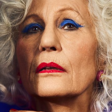 Close-up of an elderly woman with a cinematic look and warm lighting: The woman faces the camera directly, her expression both determined and challenging. Her silver-gray, curly hair frames her face. Striking stage makeup draws attention—vivid royal blue eyeshadow accentuates her eyes, while her lips are painted a bold red. Her skin shows fine lines and wrinkles, adding character and expressiveness to her face. Part of her hand is visible in the foreground and slightly out of focus, giving the image additional depth. The color palette is high-contrast and theatrical, making the figure appear strongly staged and highly present. The scene evokes the character of Rachel from Hans Werner Henze’s opera La Piccola Cubana.