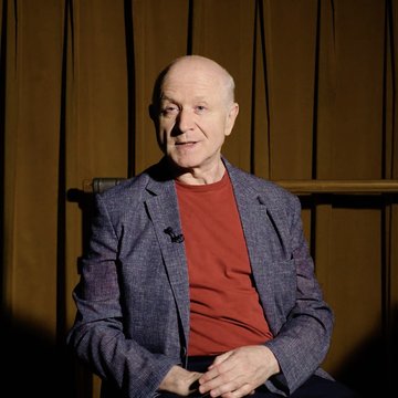 Ein älterer, glatzköpfiger Mann sitzt vor braunen Bühnenvorhängen. Er trägt ein blau strukturiertes Sakko über einem roten Shirt und scheint in einem Interview oder bei einer Präsentation zu sprechen. Warmes Bühnenlicht beleuchtet sein Gesicht und den Oberkörper. An seinem Jackett ist ein kleines Mikrofon befestigt.