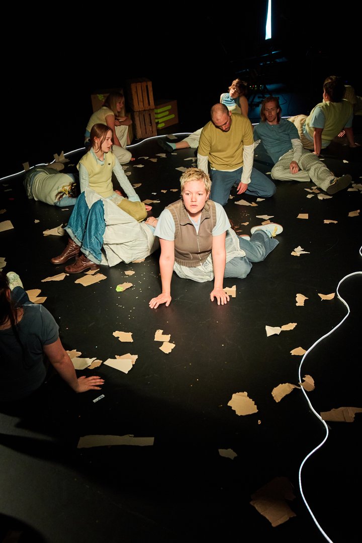 Szene aus When I find myself in Times of Trouble des Spielclubs Opera, Song and Scene. Mehrere Mitwirkende sind über die gesamte Bühne verteilt und befinden sich in unterschiedlichen Positionen auf dem Boden. Im Zentrum stützt sich eine Person mit beiden Händen auf den Boden und blickt nach vorn. Um sie herum sitzen, knien oder liegen weitere Personen, einige wirken erschöpft oder in Gedanken versunken, andere schauen zu Boden oder in die Ferne. Die Bühne ist mit zahlreichen zerrissenen Papierstücken übersät, die wie Spuren eines vorangegangenen Geschehens wirken. Ein Lichtband markiert die Grenzen des Spielraums, während Holzkisten und technische Elemente des Bühnenbilds im Hintergrund sichtbar sind. Das warme Licht hebt einzelne Figuren hervor, während große Teile des Raumes im Dunkeln bleiben.