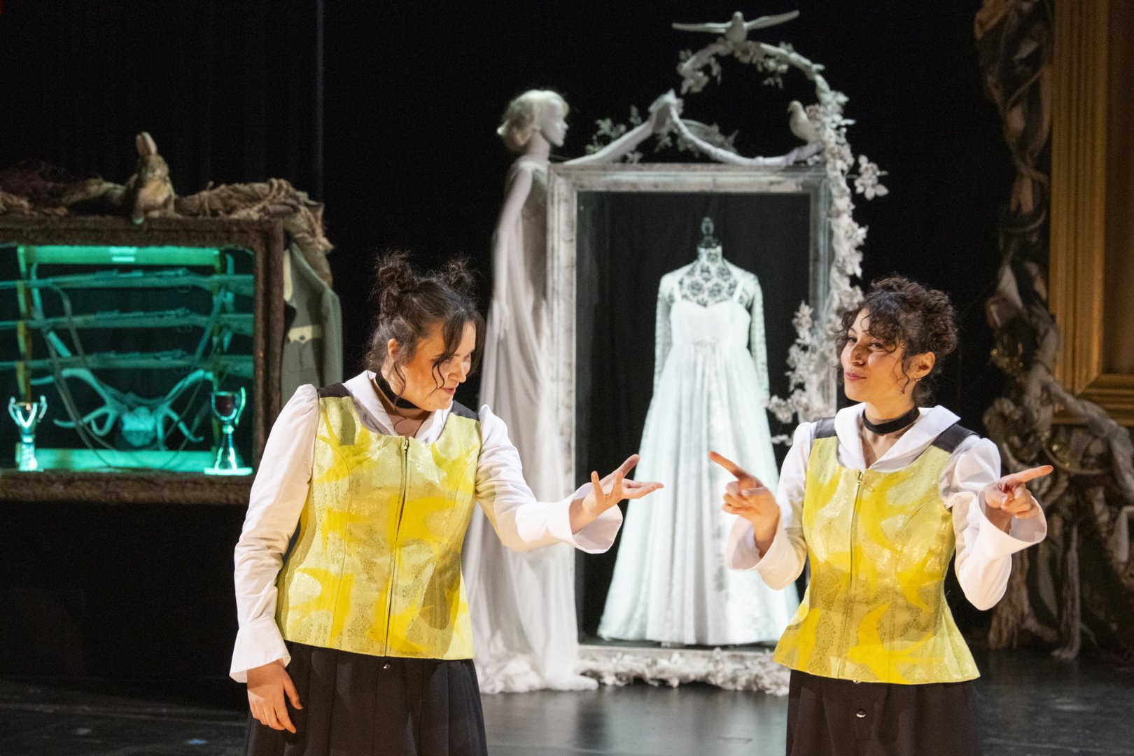 Auf einer Theaterbühne stehen zwei Darstellerinnen der Figur Agathe nebeneinander und wenden sich einander zu. Sie tragen identische Kostüme: schwarze Röcke, weiße Blusen und gelb gemusterte Westen sowie dunkle Halsbänder. Beide haben dunkles, hochgestecktes Haar. Ihre Gesten sind aufeinander bezogen und wirken wie ein Spiegelspiel; die Darstellerin rechts verwendet dabei Deutsche Gebärdensprache (DGS), wodurch die Szene als zweisprachige Darstellung lesbar wird.
Im Bühnenbild hinter ihnen steht zentral ein großer, verzierter Rahmen, in dem ein weißes Kleid wie ausgestellt präsentiert wird. Um den Rahmen herum befinden sich dekorative Elemente wie florale Ornamente und Figuren. Links im Hintergrund ist ein beleuchtetes Regal mit Requisiten zu sehen, das in grünlichem Licht schimmert. Die gesamte Szene wirkt märchenhaft und leicht surreal und stammt aus der Oper Freikugeln, in der Agathe in doppelter Ausführung gezeigt wird.