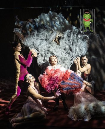 Dschilli Bey sowie Tänzer der Oper Köln in einer Szene aus der Operette Eine Frau von Format von Michael Krasznay-Krausz: Im Zentrum steht Dschilli Bey in einem auffälligen Kostüm mit üppigen roten und rosa Federapplikationen, selbstbewusst posierend und von mehreren Tänzern umgeben. Diese rahmen sie in einer stilisierten, revuehaften Choreografie ein, halten große, graue Federfächer und tragen glamouröse, teilweise transparente oder körperbetonte Kostüme. Die Szene ist in dramatisches Bühnenlicht getaucht, das mit Schatten und Lichtreflexen spielt und eine sinnlich-extravagante Atmosphäre erzeugt. Oben recht ist das Logo des Operettenfroschs, ein tanzender Frosch, von BR-Klassik platziert, den die Produktion 2025 erhielt.