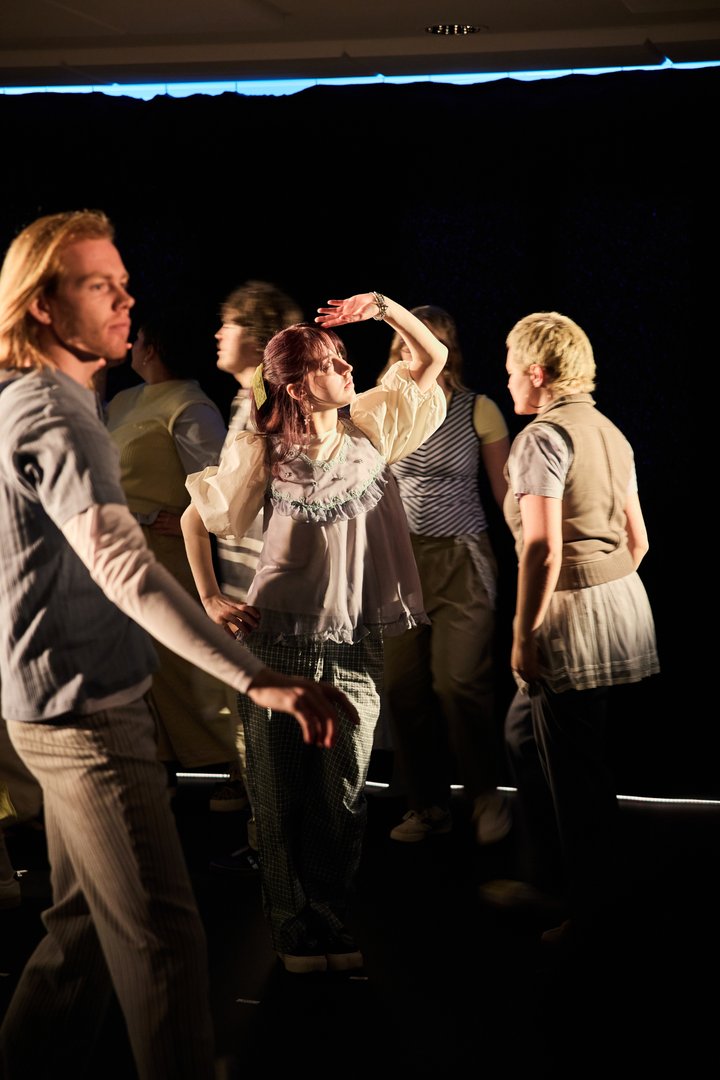 Szene aus When I find myself in Times of Trouble des Spielclubs Opera, Song and Scene. Mehrere Mitwirkende bewegen sich über eine dunkel gehaltene Bühne, die von einzelnen warmen Lichtkegeln durchbrochen wird. Im Zentrum steht eine Person mit rotbraunen Haaren, die eine Hand schützend über die Augen legt, als suche sie Orientierung oder halte Ausschau nach etwas in der Ferne. Das Licht fällt direkt auf ihr Gesicht. Um sie herum gehen andere Teilnehmende in unterschiedliche Richtungen, einige unscharf oder nur teilweise sichtbar. Ihre Bewegungen erzeugen den Eindruck von Vorbeiziehen, Suchen und Veränderung.