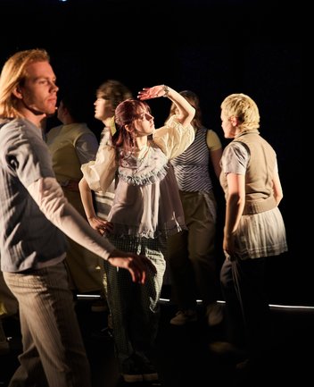 Szene aus When I find myself in Times of Trouble des Spielclubs Opera, Song and Scene. Mehrere Mitwirkende bewegen sich über eine dunkel gehaltene Bühne, die von einzelnen warmen Lichtkegeln durchbrochen wird. Im Zentrum steht eine Person mit rotbraunen Haaren, die eine Hand schützend über die Augen legt, als suche sie Orientierung oder halte Ausschau nach etwas in der Ferne. Das Licht fällt direkt auf ihr Gesicht. Um sie herum gehen andere Teilnehmende in unterschiedliche Richtungen, einige unscharf oder nur teilweise sichtbar. Ihre Bewegungen erzeugen den Eindruck von Vorbeiziehen, Suchen und Veränderung.