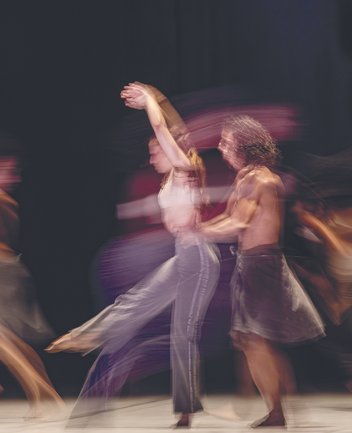 Mehrere Tänzer*innen in fließender Bewegung auf einer Bühne; im Zentrum ein Duo, das sich dynamisch dreht und hebt, mit bewusst eingesetzter Bewegungsunschärfe, aus der Choreografie „Reversible“ von Annabelle Lopez Ochoa.