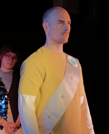 Eine Person steht frontal auf einer dunklen Bühne und blickt konzentriert nach vorne. Er trägt ein gelbes Oberteil über einem weißen Langarmshirt sowie eine schräg verlaufende Schärpe mit einem runden Emblem. Kleine Glitzerelemente schmücken das Gesicht. Im Hintergrund ist eine weitere Person unscharf zu sehen. Die Szene wirkt feierlich und performativ. Das Foto zeigt eine Szene aus der Performance „When I find myself in Times of Trouble“ des Spielclubs „Opera, Song and Scene“ der Musiktheaterpädagogik Oper.Stadt.Schule der Oper Köln.