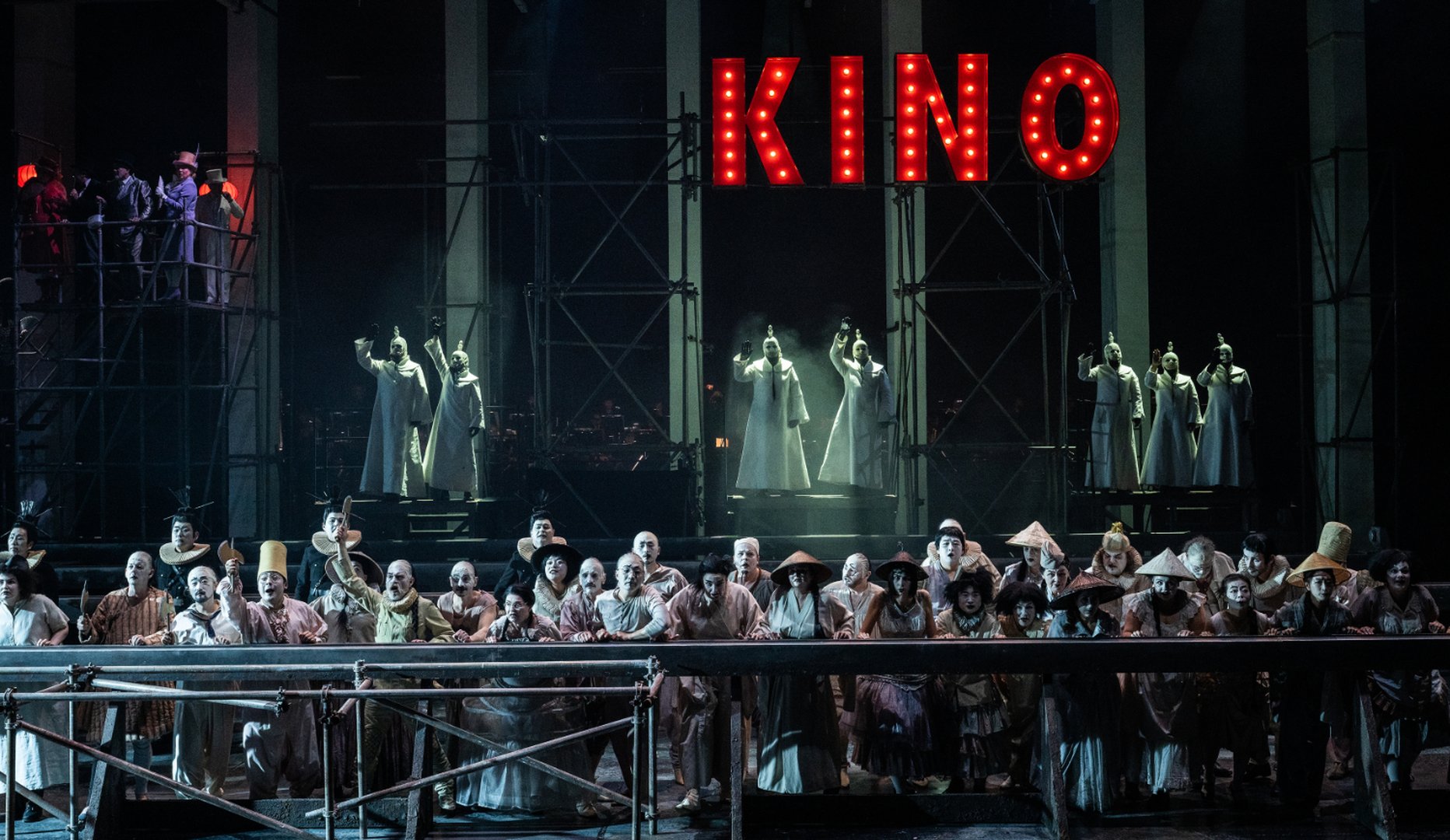Der Chor der Oper Köln steht in einer Szene aus „Turandot“ auf einer mehrstöckigen Bühnenkonstruktion. Über der Bühne leuchtet ein großes rotes „KINO“-Schild mit Glühbirnen. Die Sänger*innen tragen unterschiedliche Kostüme und blicken gespannt nach vorne. Im Hintergrund stehen mehrere Figuren in weißen Gewändern auf erhöhten Plattformen, während links oben weitere Darsteller*innen auf einem Gerüst agieren. Die Inszenierung verbindet düstere Lichtstimmung mit Elementen eines historischen Filmtheaters.