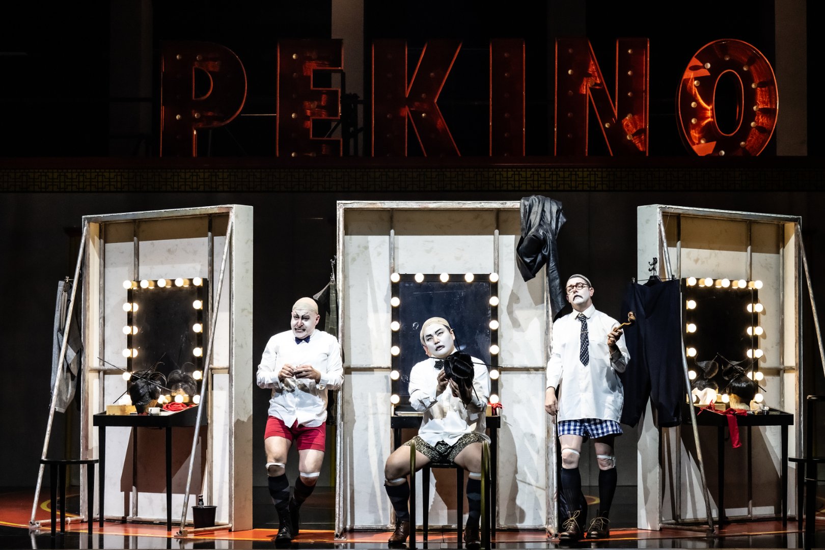 Zu sehen sind Pang, Ping und Pong in einer humorvoll inszenierten Szene aus „Turandot“. Die drei Figuren stehen vor beleuchteten Garderobenspiegeln unter einem großen „PEKINO“-Schriftzug. In Hemden, Kniestrümpfen und Unterwäsche wirken sie wie Künstler hinter der Bühne während einer Verwandlung oder Pause. Schminke, Perücken und Kostümteile liegen sichtbar auf den Tischen und unterstreichen den theaterhaften, selbstironischen Charakter der Inszenierung.