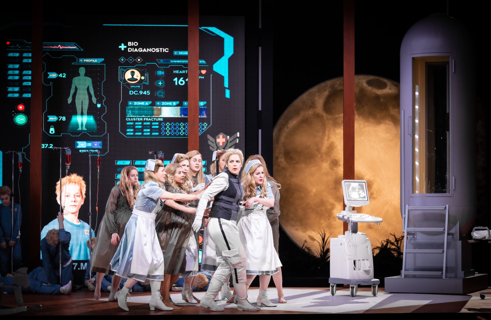 Auf einer futuristisch gestalteten Bühne versammeln sich die Walküren –Helmwige, Gerhilde, Ortlinde, Waltraute, Siegrune, Roßweiße, Grimgerde und Schwertleite – dicht um die zentrale Figur Brünnhilde, die sie stützend umgeben. Im Hintergrund sind digitale medizinische Anzeigen, Infusionsständer und ein großer, leuchtender Mond zu sehen. Die Szene verbindet mythologische Figuren mit einer modernen, klinisch-technologischen Ästhetik aus Die Walküre.