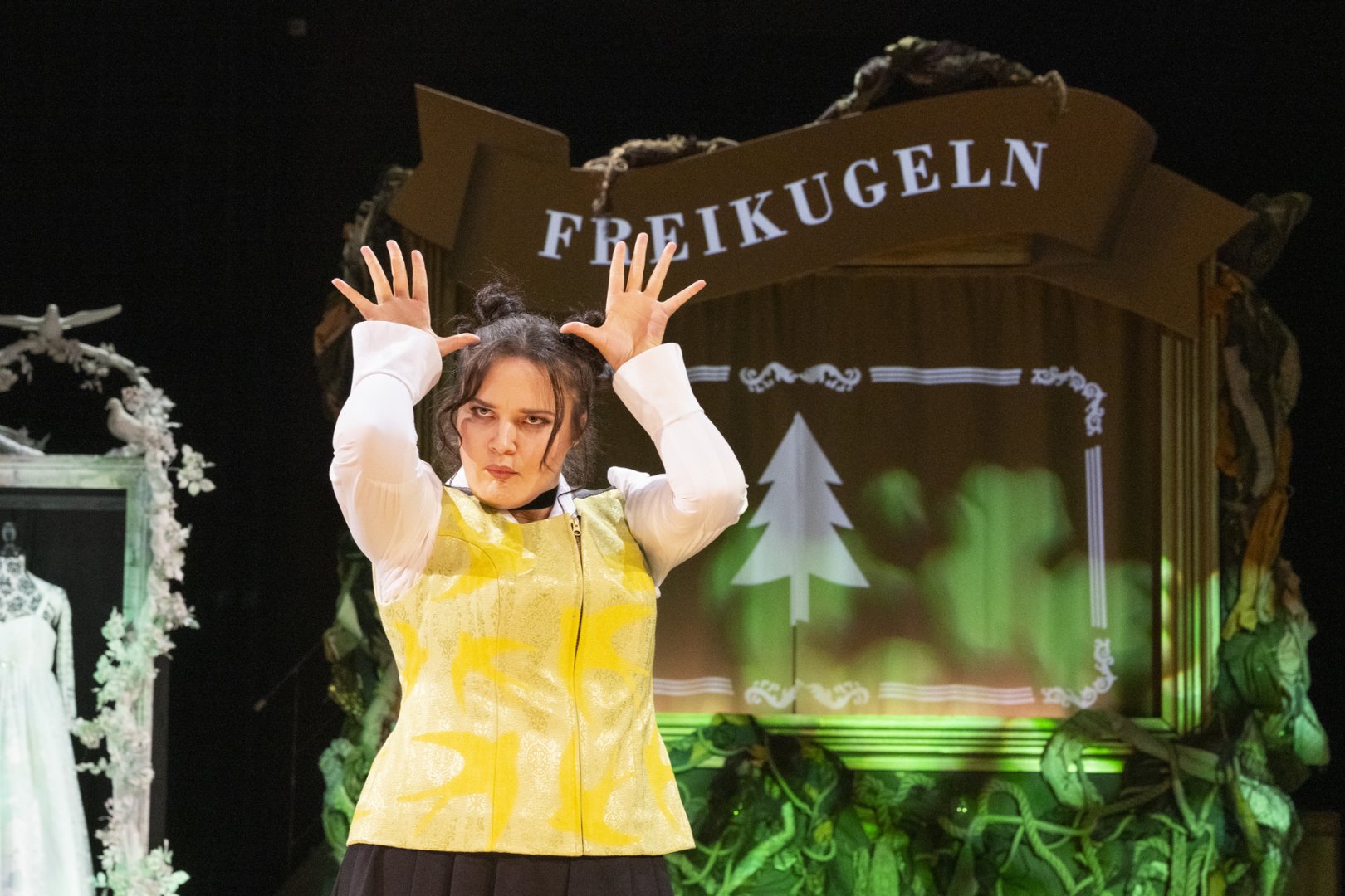Agathe aus der Oper „Freikugeln“ steht auf der Bühne vor einem dekorativen Schild mit der Aufschrift „Freikugeln“ und zeigt eine Gebärde in Deutscher Gebärdensprache. Sie trägt ein weißes Oberteil mit gelber Weste. Agathe hebt beide Hände seitlich neben ihren Kopf, etwa auf Höhe der Schläfen. Die Finger sind weit gespreizt und zeigen nach oben, die Handflächen sind nach vorne gerichtet. Durch die symmetrische Haltung beider Hände entsteht eine markante, fast kronen- oder geweihartige Form. Die Hände sind leicht nach außen gedreht, die Ellbogen zeigen ebenfalls etwas nach außen.
Ihr Gesichtsausdruck ist konzentriert und leicht angespannt, die Lippen sind zusammengepresst, was die Gebärde zusätzlich betont und ihr Ausdruckskraft verleiht. Die Haltung wirkt bewusst gesetzt und theatral, sodass die Gebärde klar sichtbar und lesbar für das Publikum ist. Im Hintergrund sind stilisierte Bühnenrequisiten und grüne Lichteffekte zu sehen.