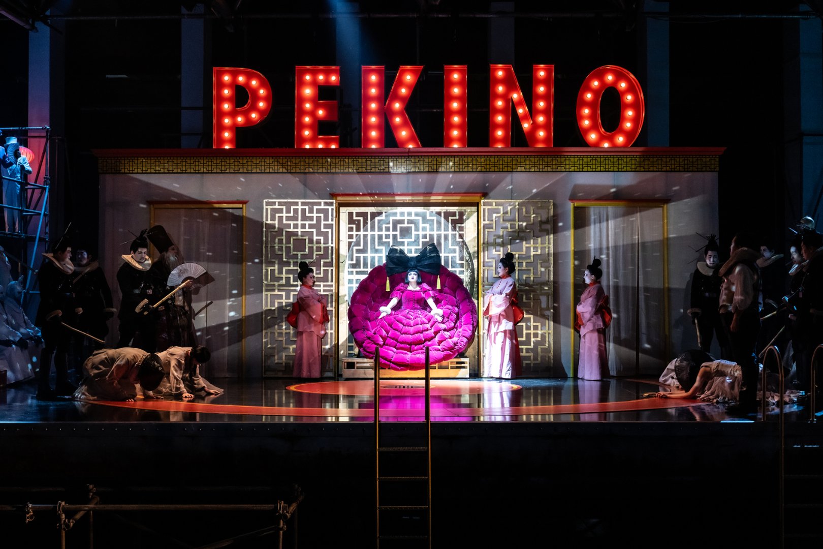 Zu sehen sind Turandot und der Chor der Oper Köln in einer opulenten Szene aus „Turandot“. Im Zentrum steht Turandot in einem auffälligen, pinkfarbenen Kleid auf einem thronartigen Sessel, flankiert von drei Dienerinnen in rosafarbenen Gewändern. Über der Bühne leuchtet ein großes „PEKINO“-Schild im Stil eines alten Kinos. Rund um die Szene stehen Mitglieder des Chors in dunklen und traditionellen Kostümen, einige kniend oder verbeugt. Die Inszenierung verbindet Elemente asiatischer Ornamentik mit glamouröser Theaterästhetik und starkem Lichtdesign.