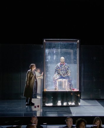Eine Frau steht links vor einer beleuchteten Glasvitrine und blickt zu einem Mann, der darin auf einem Stuhl sitzt. Die Szene ist dunkel gehalten, nur die Vitrine und die Figuren sind hell ausgeleuchtet. Im Vordergrund ist der Orchestergraben zu sehen. Die Aufnahme zeigt Woman und Artisan aus der Oper Picture a day like this.