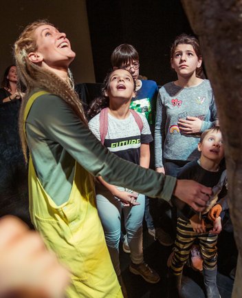 Während einer Familienführung der Oper Köln steht eine Gruppe von Kindern gemeinsam mit einer Vermittlerin auf einer Bühne. Die Vermittlerin trägt einen leuchtend gelben Overall und blickt mit einem begeisterten Ausdruck nach oben. Die Kinder schauen ebenfalls aufmerksam in dieselbe Richtung und wirken neugierig und fasziniert. Im Vordergrund rechts ist ein großes Bühnenobjekt oder Requisit angeschnitten zu sehen, das offenbar ihre Aufmerksamkeit auf sich zieht. Die warme Bühnenbeleuchtung schafft eine lebendige Atmosphäre und vermittelt den Eindruck eines gemeinsamen Entdeckens hinter den Kulissen der Oper.
