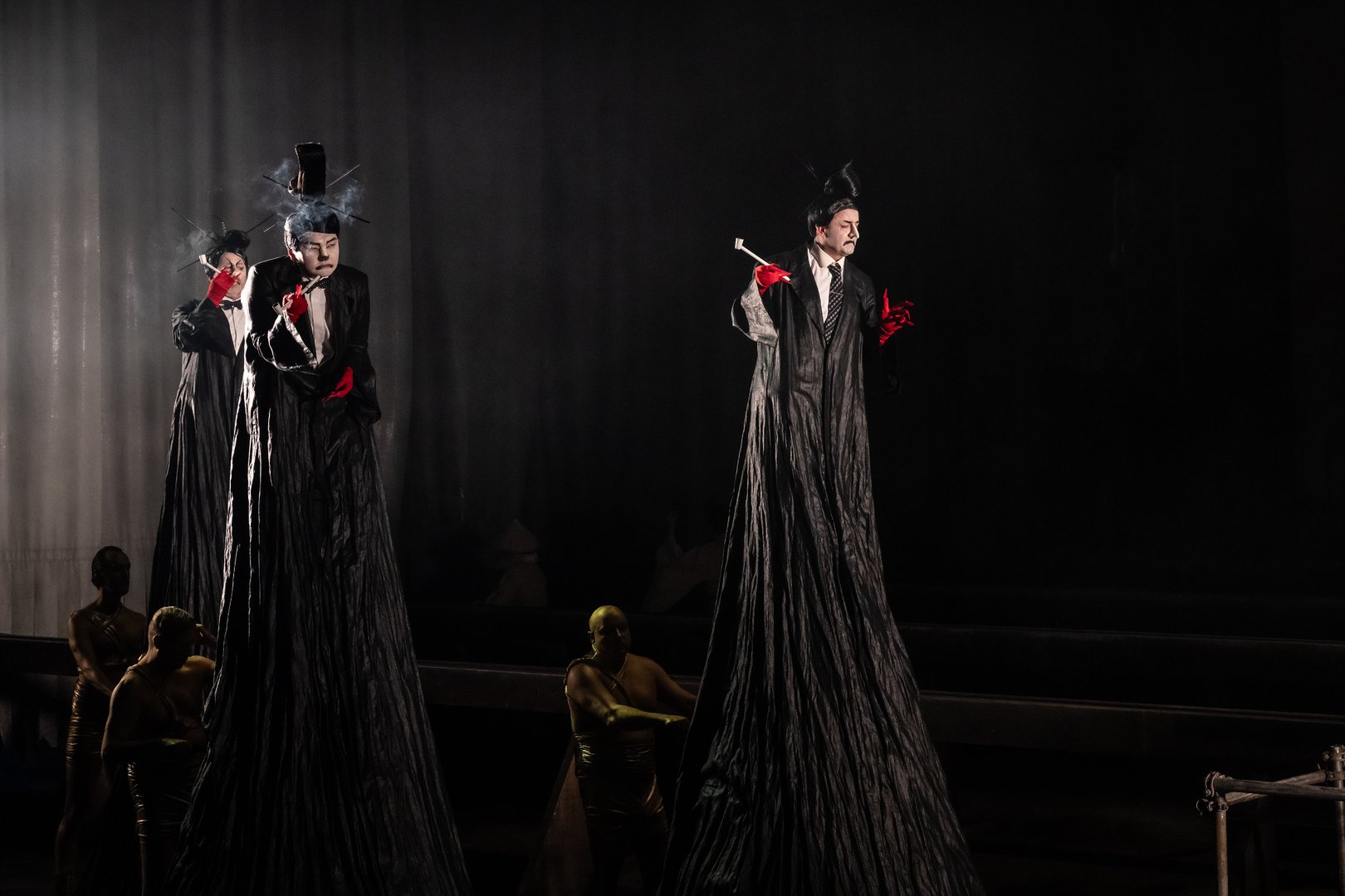 Zu sehen sind Pang, Ping und Pong sowie Mitglieder der Statistenin einer stilisierten Szene aus „Turandot“. Die drei Figuren tragen lange schwarze Gewänder auf Stelzen, weiße geschminkte Gesichter und rote Handschuhe. Mit Zigarettenspitzen und exzentrischen Frisuren wirken sie wie groteske, kabarettartige Erscheinungen. Das dunkle Bühnenbild und das gerichtete Licht verstärken die geheimnisvolle und surreale Atmosphäre der Inszenierung.