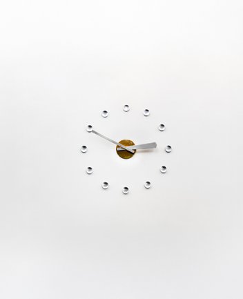 Minimalistische Wanduhr in der Oper am Offenbachplatz mit silbernen Zeigern, goldener Mitte und zwölf gläsernen Punkten als Stundenmarkierungen auf weißem Hintergrund.