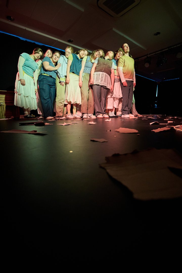 Szene aus When I find myself in Times of Trouble des Spielclubs Opera, Song and Scene. Eine Gruppe von 17 Mitwirkenden steht eng beieinander auf einer dunkel gehaltenen Bühne. Die Personen lehnen ihre Köpfe und Schultern aneinander, sodass eine dichte Formation entsteht, die wie ein einziger gemeinsamer Körper wirkt. Einige haben die Augen geschlossen, andere blicken gedankenverloren ins Leere. Die Körperhaltungen vermitteln Müdigkeit, Nachdenklichkeit und das Bedürfnis nach gegenseitigem Halt. Die Gruppe ist in sanftes, warmes Bühnenlicht getaucht, während der Hintergrund weitgehend dunkel bleibt. Auf dem Boden liegen verstreute Papierfetzen, die sich bis in den Vordergrund des Bildes ziehen und Spuren eines vorangegangenen Geschehens sichtbar machen.