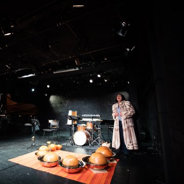 KS Dalia Schaechter in einem langen Pelzmantel steht rechts auf einer Bühne und singt mit geöffnetem Mund. Links begleiten sie zwei Musikerinnen am Flügel und Akkordeon. In der Bühnenmitte stehen ein Schlagzeug und Notenständer, im Vordergrund liegen mehrere halbkugelförmige Percussion-Instrumente auf einem Teppich. Die Szene ist in dunklem Bühnenlicht gehalten und wirkt wie eine kammermusikalische Theaterperformance. Links im HIntergurnd sitzen Dorrit Bauerecker (Akkordeon), Theresia Renelt (Klavier) und  Kurt Fuhrmann (Schlagzeug).