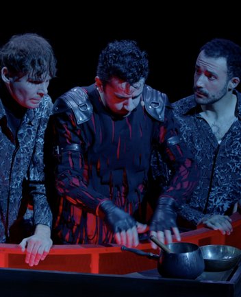 Drei junge Männer stehen auf einer Bühne in einem runden, rot leuchtenden Becken, das die Wolfsschlucht darstellt. In der Mitte befindet sich Kaspar in dunkler, markanter Kleidung und gießt konzentriert Freikugeln in einem kleinen Topf – ein zentraler Moment aus der Oper „Der Freischütz“ von Carl Maria von Weber. Rechts und links von ihm stehen zwei Darsteller der Figur Max – ein Sänger und ein DGS-Schauspieler – und beobachten die Szene angespannt. Die Beleuchtung in intensiven Rot- und Blautönen verstärkt die düstere, unheilvolle Atmosphäre.