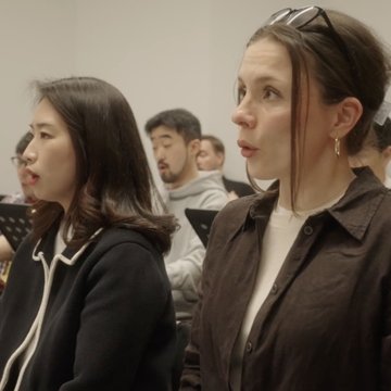 Probe des Chors der Oper Köln: Mehrere Sängerinnen und Sänger sitzen im Probenraum, konzentriert auf ihre Notenblätter, und singen gemeinsam eine Passage. Im Vordergrund sind zwei Frauen im Profil zu sehen, während im Hintergrund weitere Chormitglieder aufmerksam folgen – eine Momentaufnahme intensiver musikalischer Arbeit.