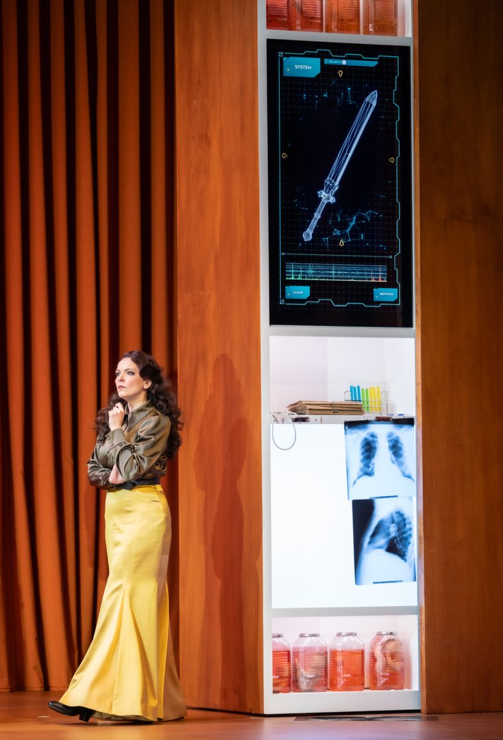 Auf einer modern gestalteten Bühne steht Fricka nachdenklich mit verschränkten Armen vor einem warm orangefarbenen Vorhang. Neben ihr ist ein futuristisch wirkendes Wandmodul zu sehen, das eine digitale Darstellung eines Schwerts sowie medizinische Aufnahmen zeigt. Die Szene verbindet klassische Opernfigur mit einer zeitgenössischen, technologischen Ästhetik aus Die Walküre.