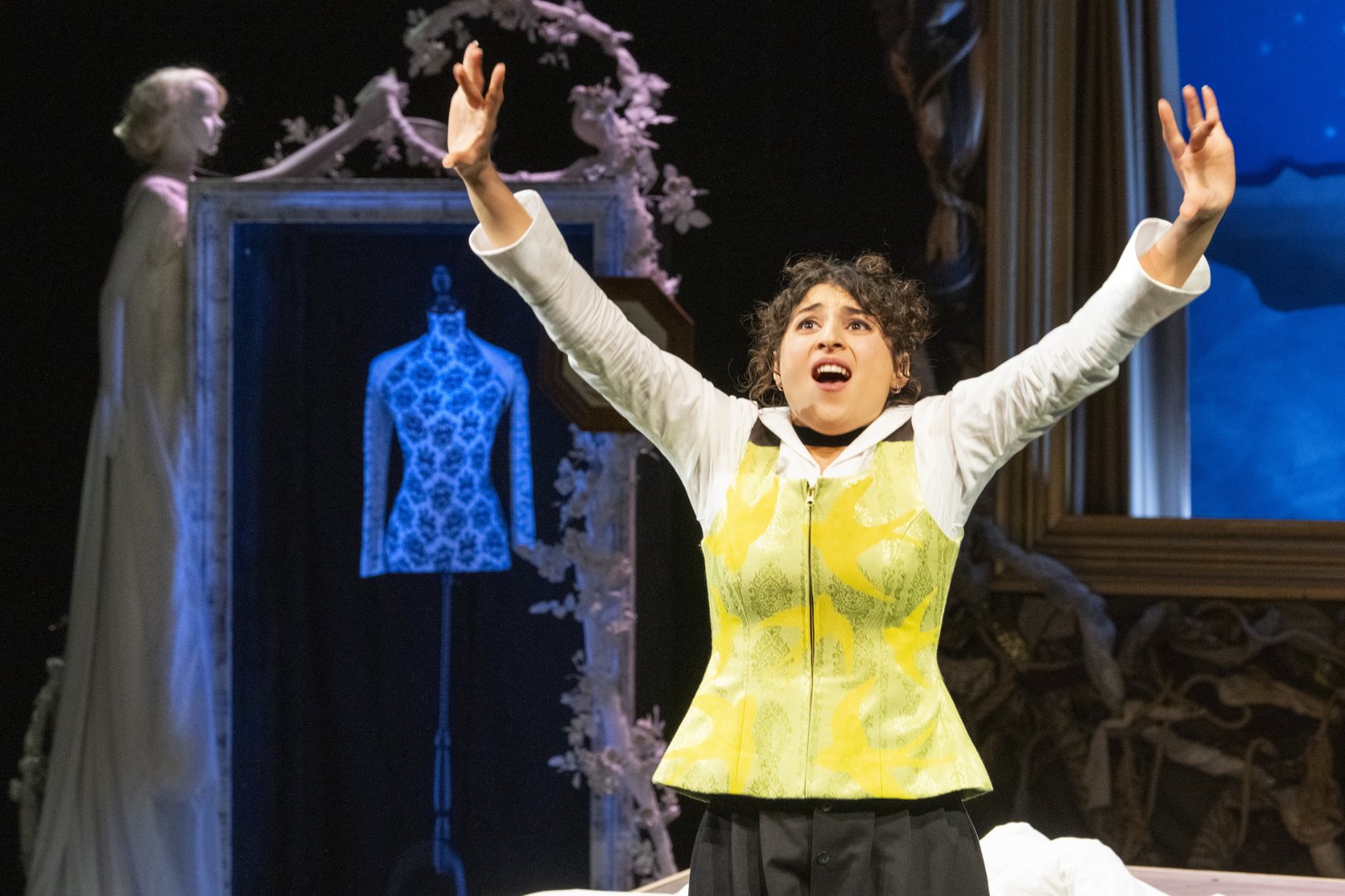 Auf einer Theaterbühne ist eine Darstellerin als Agathe aus der Oper „Freikugeln“ zu sehen. Sie steht im Vordergrund, hebt beide Arme weit nach oben und öffnet den Mund, als würde sie kraftvoll singen. Ihr Gesichtsausdruck ist intensiv und emotional, zwischen Hoffnung und Anspannung.
Sie trägt ein weißes Hemd mit einer auffälligen, gelb-grün gemusterten Weste sowie dunkle Hosen. Ihr lockiges Haar ist locker hochgesteckt. Die Körperhaltung ist aufgerichtet und ausdrucksstark, wodurch die Szene eine starke dramatische Wirkung erhält.
Im Hintergrund ist ein dekorativer Bühnenraum zu sehen: Ein großes, rahmenartiges Element zeigt eine Schneiderpuppe mit einem blau leuchtenden Oberteil. Daneben steht eine helle, skulpturartige Frauenfigur, umgeben von einem floralen, kreisförmigen Ornament mit weißen Blüten und kleinen Vogelelementen. Rechts ist ein weiterer gerahmter Bereich mit kühlem, blauem Licht angedeutet.
Die Lichtgestaltung kombiniert warme und kühle Töne und verstärkt die emotionale Spannung der Szene zwischen innerem Aufruhr und märchenhafter, leicht entrückter Atmosphäre.