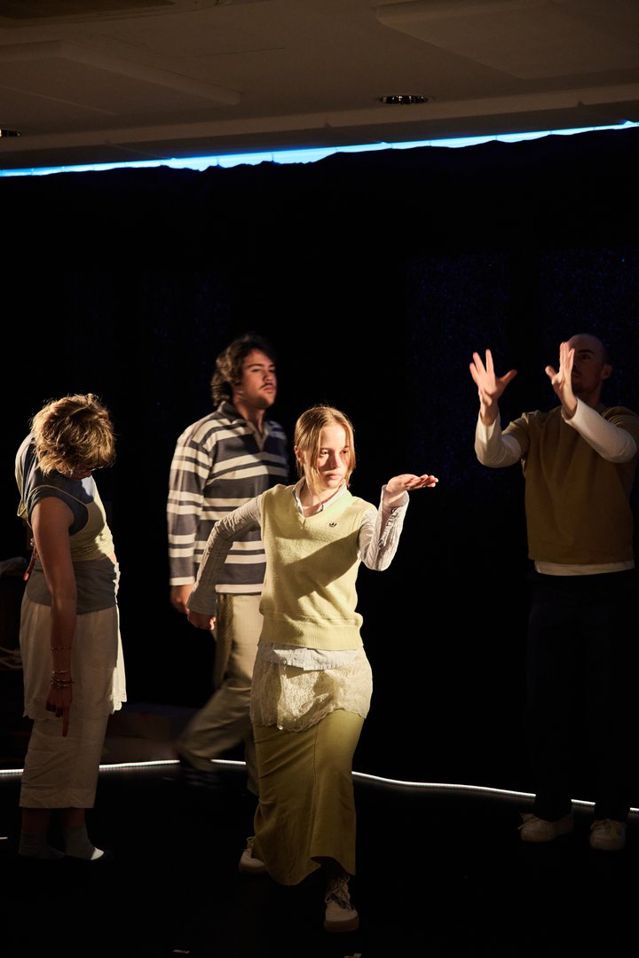 Szene aus When I find myself in Times of Trouble des Spielclubs Opera, Song and Scene. Auf einer dunkel gehaltenen Bühne stehen vier Mitwirkende in unterschiedlichen Posen, die durch gezielte Lichtinseln hervorgehoben werden. Im Zentrum geht eine junge Person in einem hellen Rock und einem gelben Pullunder in die Knie. Mit konzentriertem Blick hält sie eine Hand waagerecht vor sich, während die andere hinter ihrem Rücken angewinkelt ist.
Um sie herum befinden sich weitere Personen, die jeweils eigene Bewegungen ausführen: Links steht eine Person mit gesenktem Kopf und herabhängenden Armen, ihre Körperhaltung vermittelt Nachdenklichkeit oder Erschöpfung. Dahinter bewegt sich eine weitere Person durch den Raum, als sei sie auf dem Weg von einem Ort zum anderen. Rechts hebt eine Person beide Hände in die Höhe, als wolle sie etwas auffangen, erreichen oder festhalten. Zwischen den Figuren entsteht keine direkte Interaktion, dennoch sind sie durch den gemeinsamen Bühnenraum miteinander verbunden.