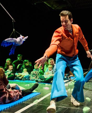 Ein Performer in orangefarbenem Oberteil und blauer Hose bewegt sich dynamisch auf der Bühne und interagiert mit Babys und Kleinkindern, die zusammen mit ihren Begleitpersonen auf Matten sitzen. Über der Szene schwebt ein spielerisches Objekt an einer Schnur. Die Kinder beobachten das Geschehen aus nächster Nähe in einer offenen, interaktiven und farbenfroh beleuchteten Bühnenatmosphäre.