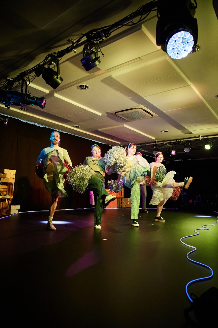 Szene aus When I find myself in Times of Trouble des Spielclubs Opera, Song and Scene. Vier Mitwirkende stehen nebeneinander auf einer offenen Bühne und führen eine synchronisierte Choreografie aus. Mit erhobenen Knien und ausgestreckten Beinen setzen sie zu einem tänzerischen Schritt an. In ihren Händen halten sie glitzernde Pompons in Silber und Rot, die das Bühnenlicht reflektieren und der Szene eine festliche, beinahe ausgelassene Atmosphäre verleihen. Die Gesichter der Darstellenden zeigen Konzentration und Freude, während sie gemeinsam in dieselbe Bewegungsrichtung agieren. Über ihnen sind Scheinwerfer und technische Elemente des Aufführungsraums sichtbar, die Bühne wird von farbigen Lichtakzenten in Grün, Blau und warmen Tönen beleuchtet.
Der Moment erinnert an eine Cheerleading- oder Revue-Nummer und hebt sich durch seine Leichtigkeit und seinen spielerischen Charakter hervor.
