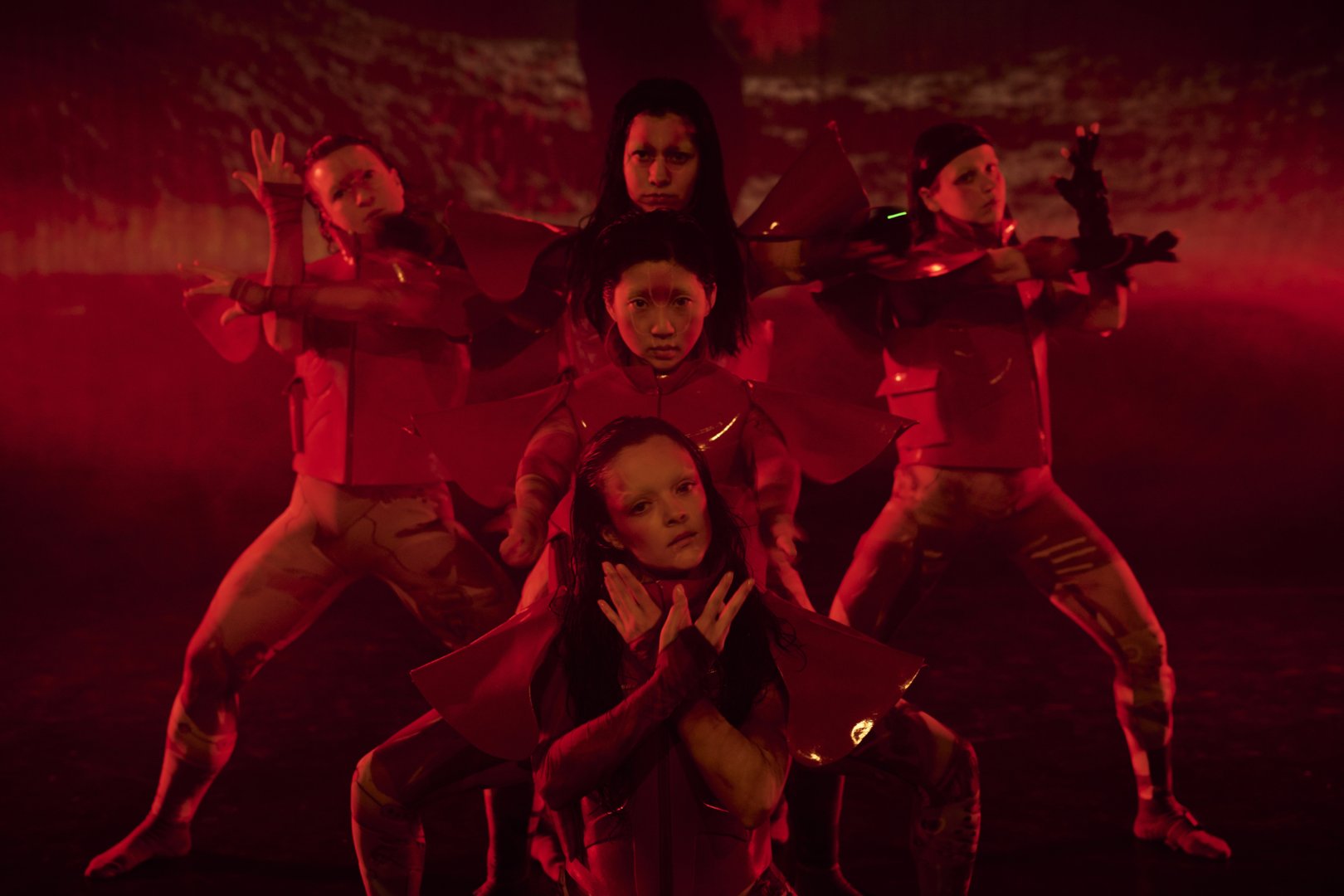 Fünf Tänzerinnen in futuristisch wirkenden Kostümen stehen in einer symmetrischen Formation auf einer intensiv rot beleuchteten Bühne. Im Vordergrund sitzt eine Person und verschränkt die Hände vor dem Hals, die Finger fächerförmig gespreizt und leicht nach oben gerichtet, sodass sie wie ein schützendes oder rahmendes Element wirken. Dahinter staffeln sich die weiteren Performerinnen, deren Arme seitlich ausgestreckt sind. Ihre Hände formen präzise, stilisierte Gesten: Finger sind einzeln angehoben, gekrümmt oder gespreizt, teilweise wie Zeichen oder Codes, die in die Luft geschrieben werden. Die Handhaltungen wirken kontrolliert und synchron, fast mechanisch, und verstärken die strenge, technologische Ästhetik der Szene. Die Gesichter sind ernst und konzentriert, die gesamte Komposition wirkt ruhig, kraftvoll und zugleich leicht unheimlich.