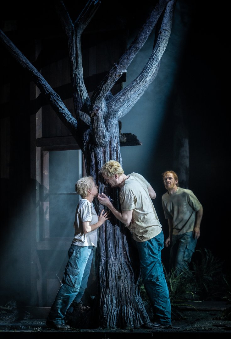 Auf einer düster beleuchteten Bühne mit einem großen, kahlen Baum stehen drei Figuren: Siegmund und Sieglinde lehnen sich einander intensiv zugewandt versteckt vor dem Baumstamm gegenüber, während Hunding im Hintergrund des Baumes wachsam und bedrohlich beobachtet. Die Szene ist in kühles, dramatisches Licht getaucht und erzeugt eine gespannte, emotionale Atmosphäre aus Die Walküre von Richard Wagner.