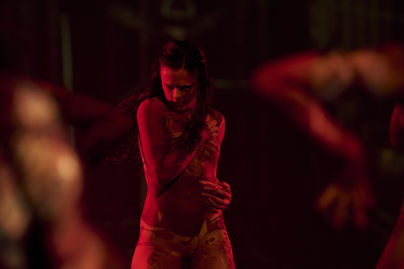 Eine Tänzerin steht allein im Zentrum einer rot ausgeleuchteten Bühne. Ihr Körper ist leicht eingedreht, der Blick konzentriert und nach unten gerichtet. Eine Hand liegt fest an ihrer Schulter, während die andere den Unterbauch umfasst – die Finger greifen dabei leicht ein, als würde sie sich selbst halten oder schützen. Die Hände wirken angespannt und zugleich kontrolliert, mit deutlich sichtbarer Muskelspannung. Unscharfe Arme anderer Performer*innen rahmen die Szene im Vordergrund, wodurch die Figur isoliert und innerlich fokussiert erscheint. Die Stimmung ist intensiv, introvertiert und emotional aufgeladen.