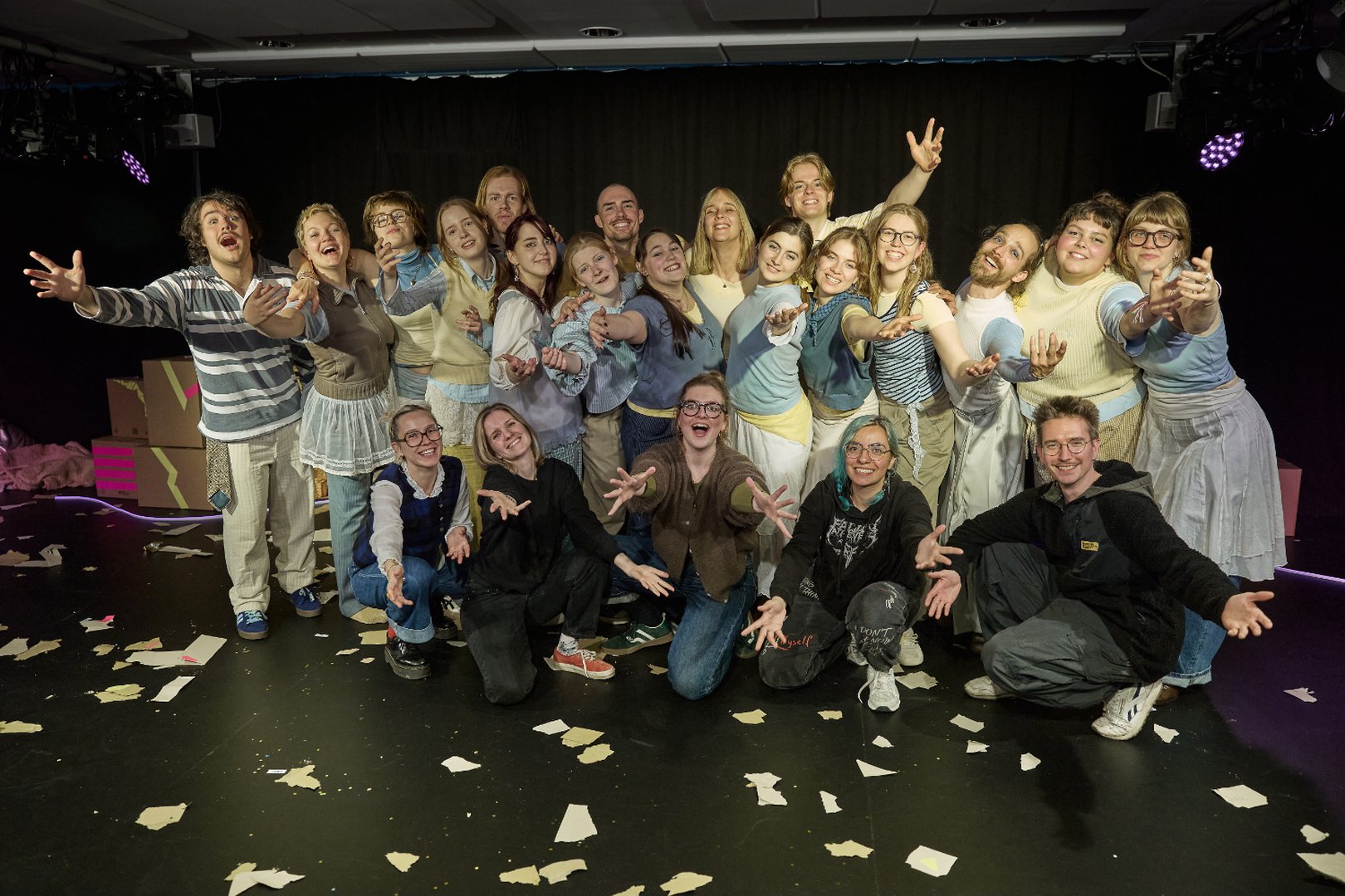 Gruppenfoto des gesamten Teams von When I find myself in Times of Trouble des Spielclubs Opera, Song and Scene. Auf der Bühne versammeln sich die Mitwirkenden der Produktion gemeinsam mit dem künstlerischen Leitungsteam zu einem Abschlussbild. Die Gruppe steht und kniet in mehreren Reihen vor einem dunklen Bühnenhintergrund. Viele Personen strecken die Arme offen in Richtung Kamera, einige lachen, andere lächeln breit oder blicken freudig und stolz in die Linse. Die lockeren Posen und die Nähe zueinander vermitteln Vertrautheit und Gemeinschaft. Auf dem Boden liegen verstreute Papierfetzen aus der Inszenierung, während Teile des Bühnenbilds mit Kisten und farbigen Markierungen im Hintergrund sichtbar bleiben.