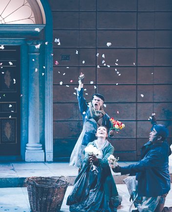 Szene aus My Fair Lady von Frederick Loewe: Drei Darstellende in historisch anmutender Kleidung befinden sich auf einer Straßenkulisse vor einer Hausfassade mit Säulenportal (Covent-Garden-Blumenmarkt): Ein älterer Mann im Hintergrund, gekleidet in Mantel, Schal und Mütze, wirft Blumen in die Luft. Im Vordergrund kniet eine junge Frau, Eliza Doolittle, in einem langen Kleid und hält einen Strauß weißer Rosen, während sie freudig nach oben blickt. Neben ihr kniet ein junger Mann in dunklem Mantel und Schirmmütze, ebenfalls mit Blumen in der Hand, und schaut zu ihr hinüber. Herabfallende Blütenblätter durchziehen die Szene und erzeugen eine lebhafte, festliche Atmosphäre, die an das geschäftige Treiben eines Blumenmarkts erinnert.