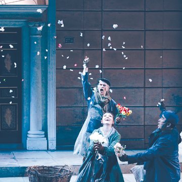 Scene from My Fair Lady by Frederick Loewe: Three performers in period-style costumes are set in a street scene in front of a building façade with a columned entrance (Covent Garden flower market). An older man in the background, wearing a coat, scarf, and cap, tosses flowers into the air. In the foreground, a young woman, Eliza Doolittle, kneels in a long dress holding a bouquet of white roses, looking up with delight. Beside her, a young man in a dark coat and flat cap also kneels, holding flowers and looking toward her. Falling petals fill the scene, creating a lively, festive atmosphere reminiscent of the bustling activity of a flower market.