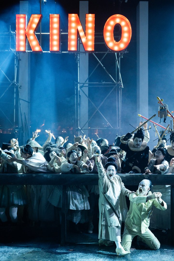 Der Chor der Oper Köln drängt sich in einer dramatischen Szene aus „Turandot“ auf die Bühne. Über den Sänger*innen leuchtet ein großes „KINO“-Schild im Stil alter Leuchtreklamen. Im Vordergrund stehen zwei Solist*innen in hellen Kostümen, gestikulierend und voller Ausdruck. Die Inszenierung wirkt turbulent und theatral, mit dichtem Bühnenbild, Nebel und starkem Lichtkontrast zwischen kaltem Blau und warmem Neonlicht.